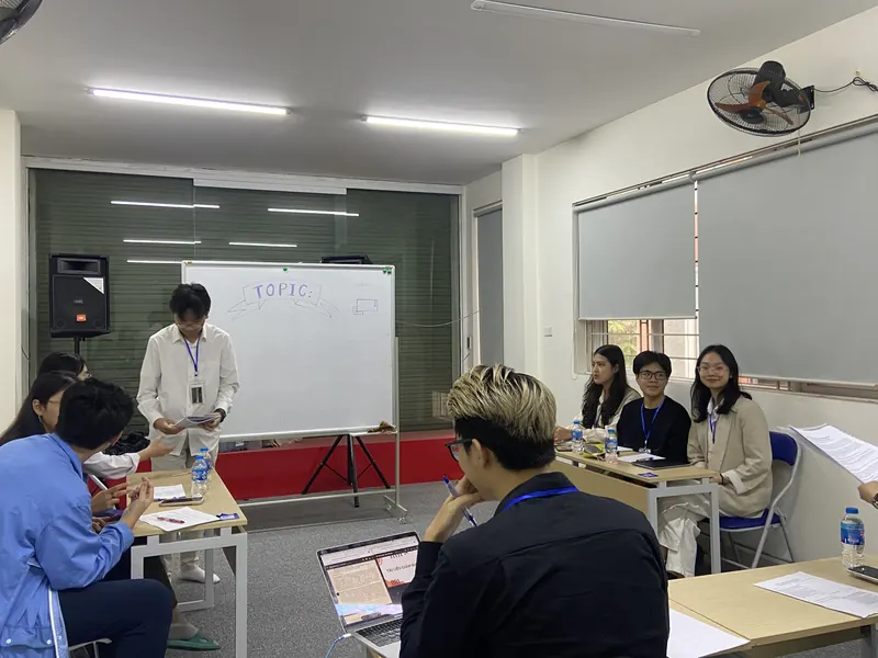 A student speaking at the front of the room while the judges score from their table.